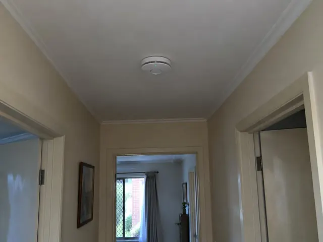 Ceiling-mounted smoke alarm in a beige hallway with doorways and crown molding in Hamlyn Terrace